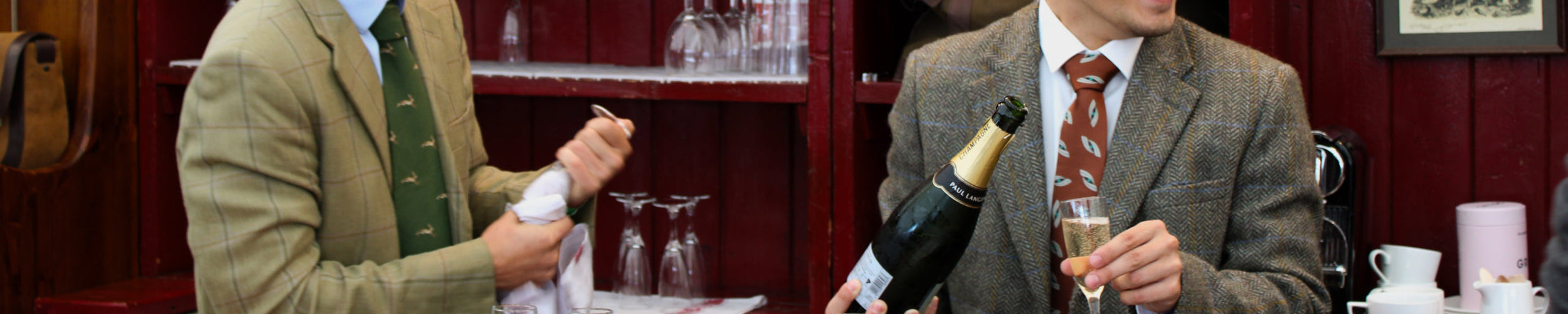 Wide image of Two men pouring champagne at the Dubarry stand at the Game Fair in 2023. Both men are wearing grey and green tweed jackets; one is wearing a green tie, while the other wears a red tie.