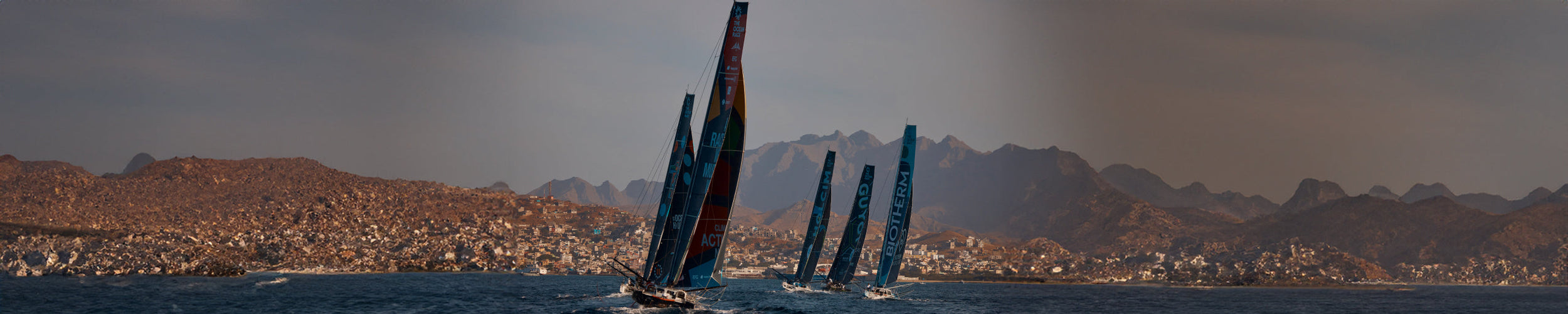 Wide image of five ships sailing at sea, with the town of Mindelo in the background, as they set off on the Ocean Race second leg 2023.