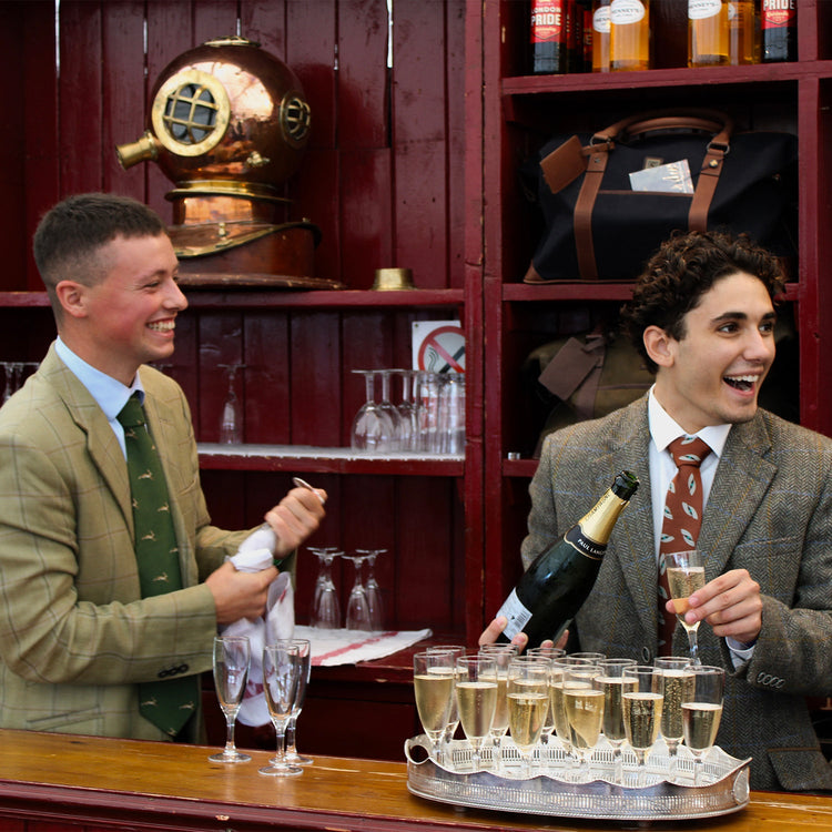 Square image of Two men pouring champagne at the Dubarry stand at the Game Fair in 2023. Both men are wearing grey and green tweed jackets; one is wearing a green tie, while the other wears a red tie.//© Dubarry of Ireland